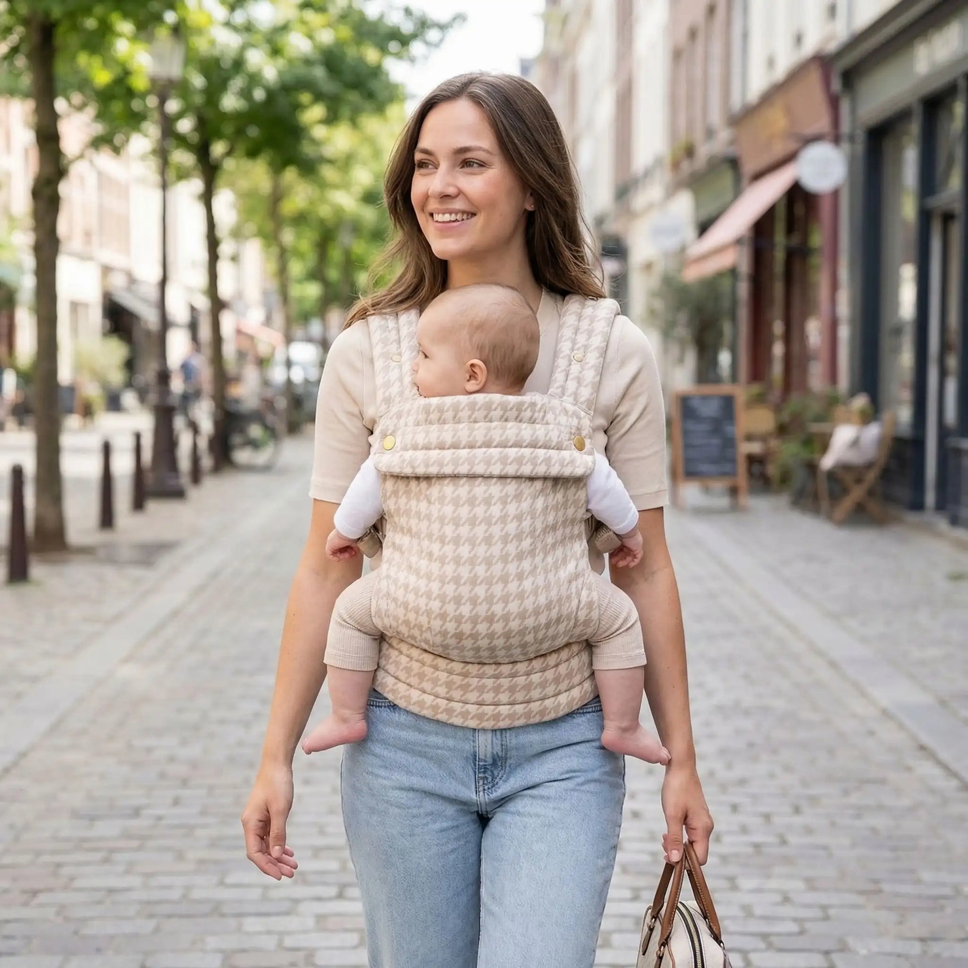 maman souriante marchant en ville avec bebe dans un porte bebe ergonomique beige
